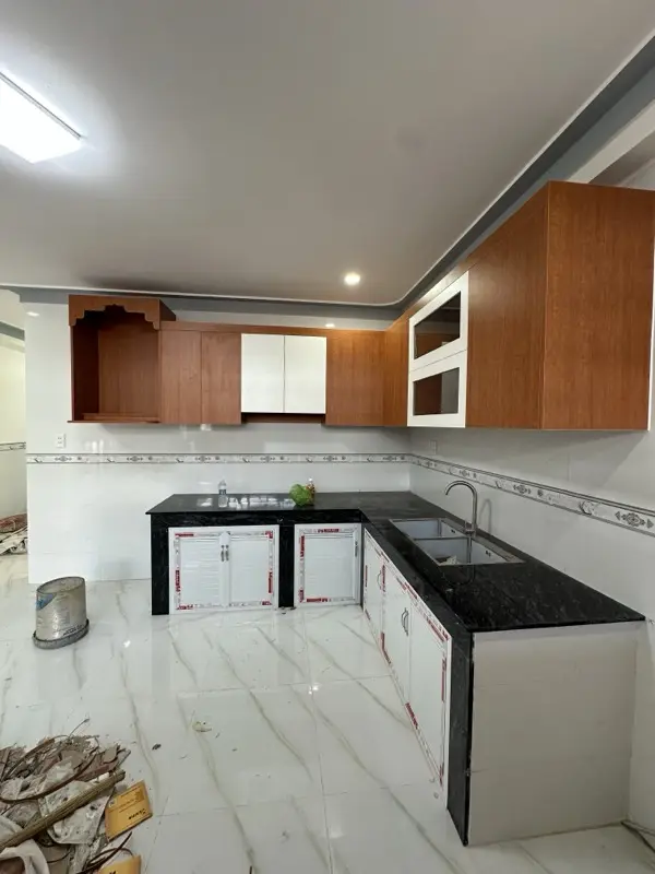 Tủ bếp chữ L vừa lắp tủ trên vân gỗ, cánh kính khung trắng, sàn còn vật tư